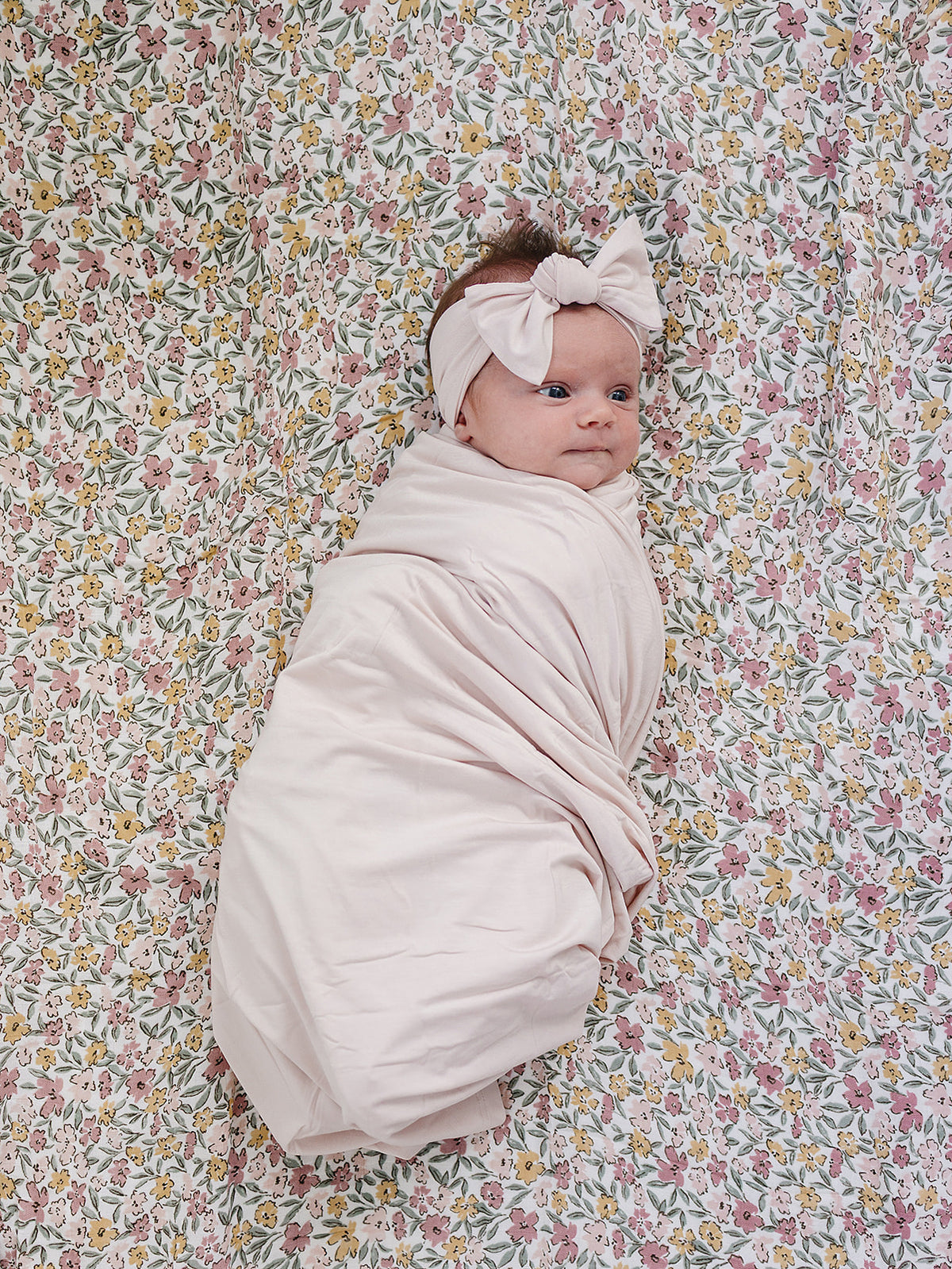 A baby swaddled in a light pink blanket and wearing a matching cream pink headband, lying on a floral fabric background.