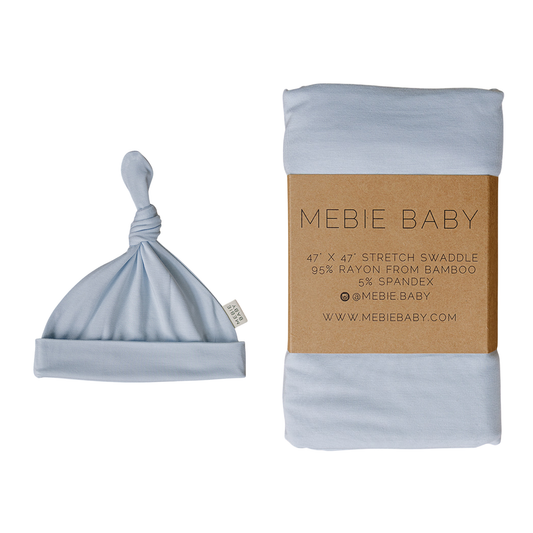 A light blue bamboo baby hat with a knot is shown next to a folded matching swaddle blanket wrapped in a brown paper band labeled 'MEBIE BABY'.