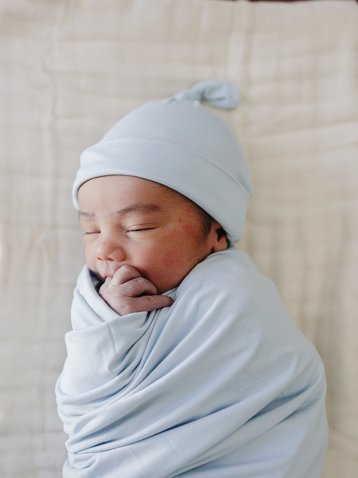 A sleeping newborn baby swaddled in a light blue wrap and wearing a matching light blue knot hat.
