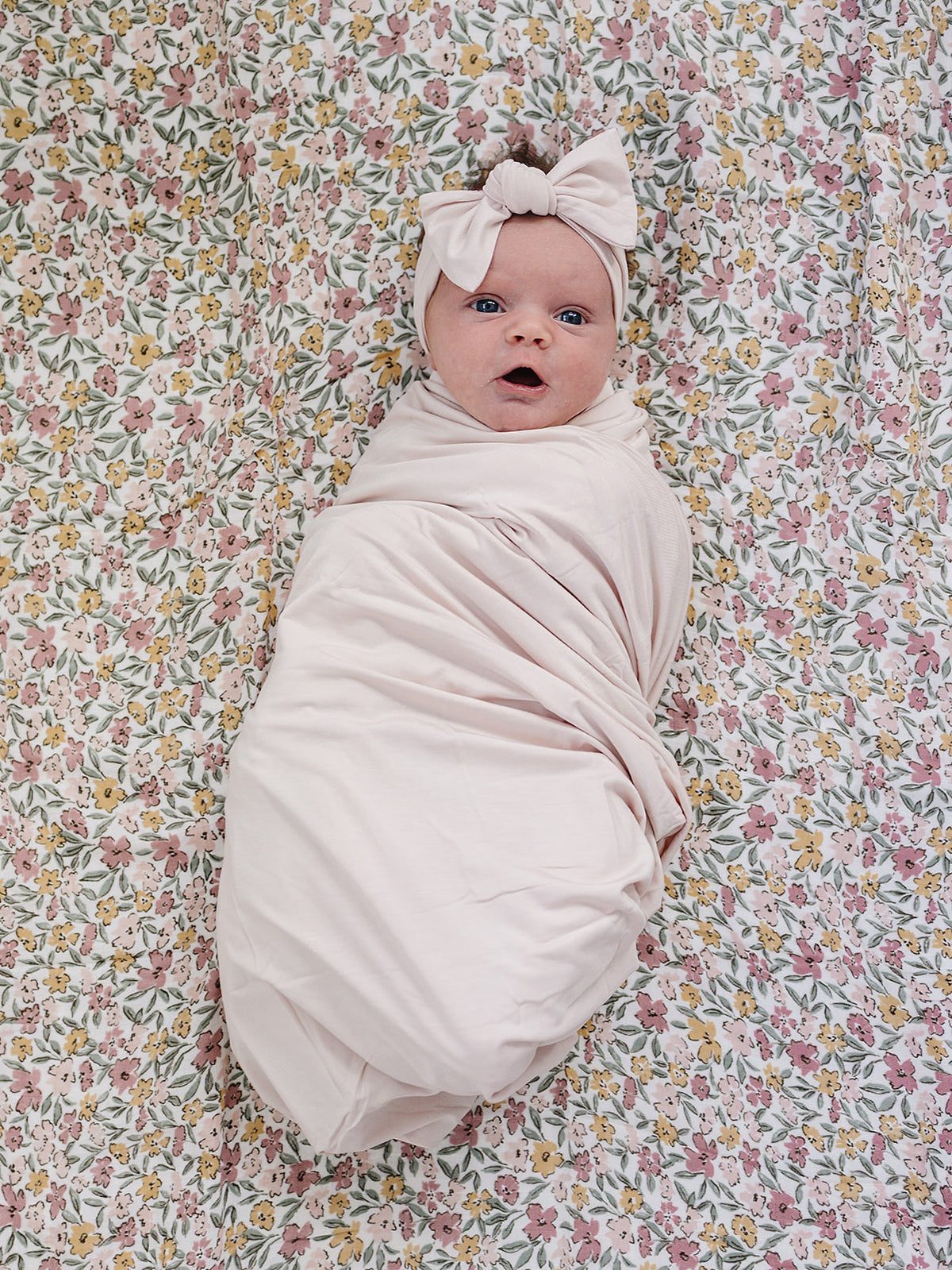 A swaddled baby lying on a floral blanket, wearing a matching pale pink bow headband. The swaddle and headband are a very light blush pink, contrasting with the multicolored floral background.
