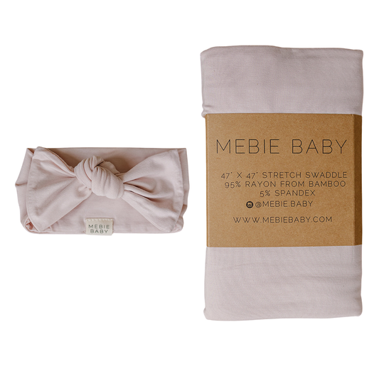 A folded baby swaddle blanket in a soft blush pink color, shown in two states: on the left as a knot head wrap. The brand 'Mebie Baby' is clearly printed on the band and a small tag on the bow.